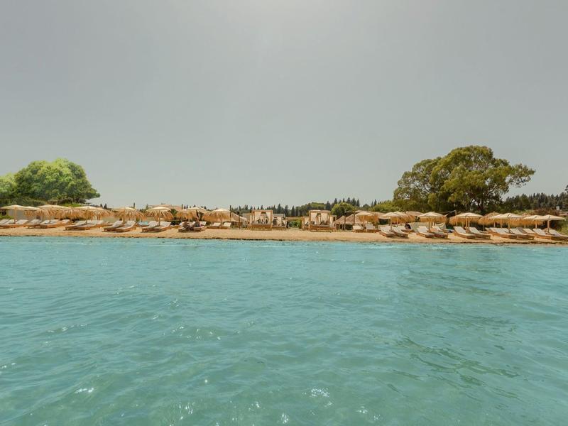 Klares blaues Meer mit felsiger Küste und vereinzelten Bäumen im Hintergrund unter hellem Himmel.