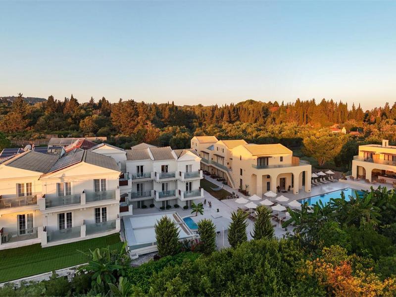 Großes Hotel mit mehreren Gebäuden, Pool und Garten, umgeben von Bäumen und klarem Himmel.