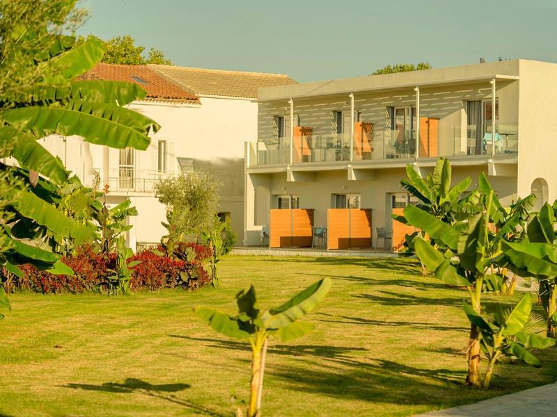 Zweistöckiges Haus mit weißen Wänden, Balkon und grünem Garten mit jungen Bananenpflanzen.