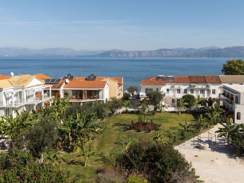 Blick auf weiße Häuser mit roten Dächern, grünen Gärten und blauem Meer im Hintergrund.