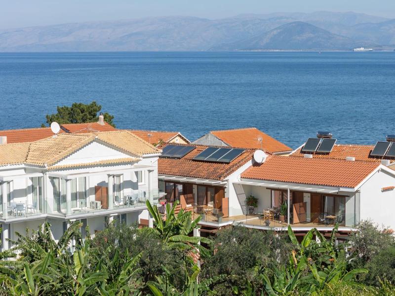 Mehrere Häuser mit roten Dächern und Solarpanelen am Meer vor Bergen, üppige Vegetation im Vordergrund.