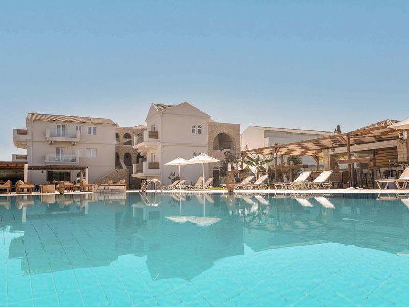 Großer klarer Pool vor weißen Hotelgebäuden und Sonnenliegen unter blauem Himmel.