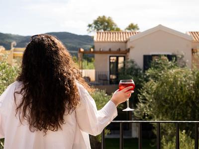 Femme aux longs cheveux tenant un verre sur un balcon avec vue sur des maisons et des montagnes.