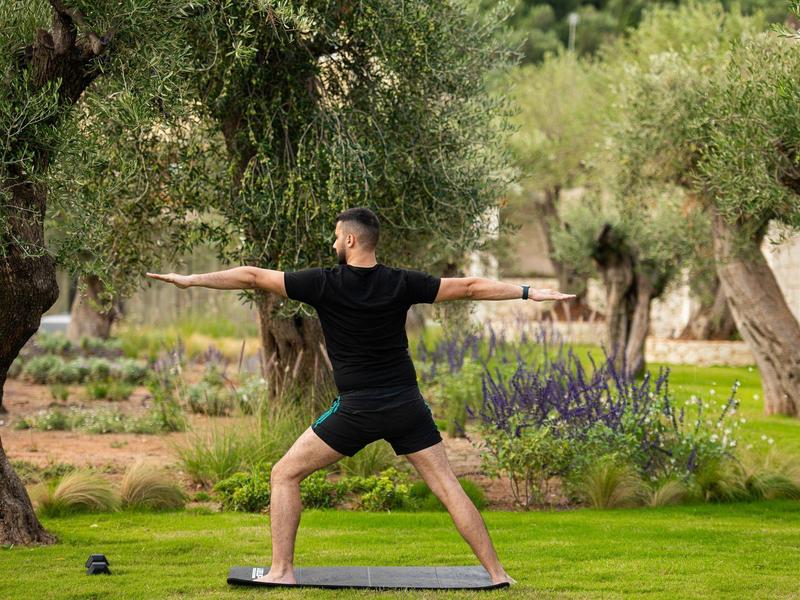 Person macht Yoga im Freien auf einer Matte umgeben von Olivenbäumen.