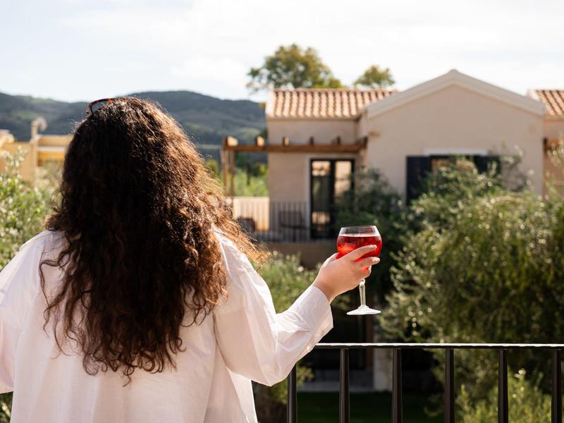 Person mit langem Haar in weißem Hemd hält ein Glas Wein auf Balkon mit Blick auf ein helles Ferienhaus.