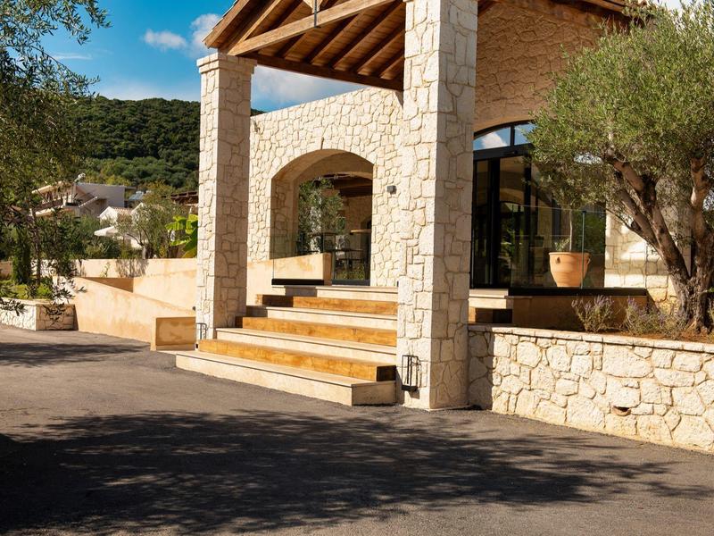 Eingang eines Steinhauses mit Stufen und großen Töpfen, umgeben von Bäumen und Sonnenschein.