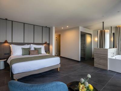 Helle, moderne Hotellzimmer mit großem Bett, grauen Fliesen und Sitzecke mit blauen Stühlen.