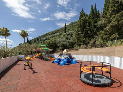 Kinderspielplatz mit Karussell, Rutsche und Klettergerüst, umgeben von grünen Hügeln und blauem Himmel.