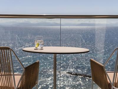 Balkontisch mit zwei Stühlen, Blick auf glitzerndes Meer und bewölkten Himmel im Hintergrund.