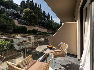 Balkon mit Glasgeländer, zwei Stühlen und kleinem runden Tisch, umgeben von grünen Hügeln.