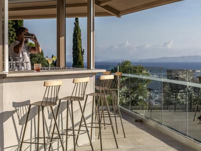 Offene Terrasse mit Bar, vier hohen Metallstühlen und Meerblick im Hintergrund bei klarem Himmel.