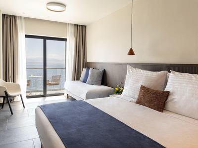 Helles Schlafzimmer mit großem Bett, grauem Sofa, weißem Stuhl und Balkon mit Meerblick.