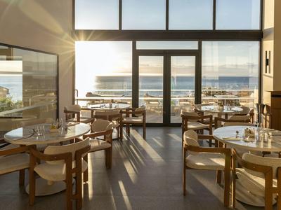 Helles Café mit Holzstühlen und Tischen, großen Fenstern und Meerblick bei Sonnenuntergang.