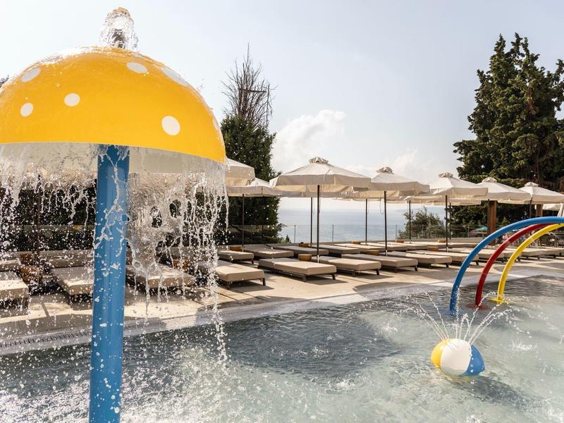 Kinderplanschbecken mit gelbem Pilz-Wasserspender und bunten Wasserbögen, Liegen im Hintergrund.