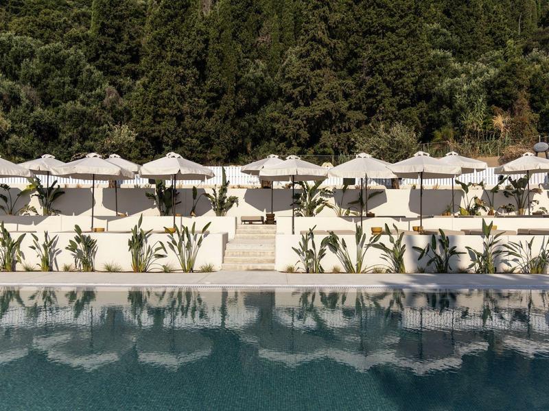 Ein klarer blauer Pool mit weißen Sonnenschirmen und Liegen davor, im Hintergrund grüne Bäume.