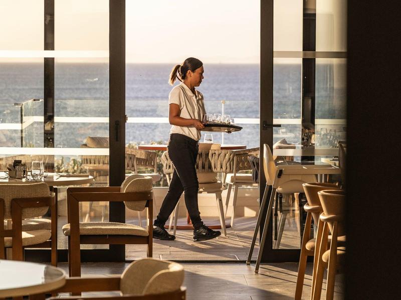 Eine Kellnerin trägt Tablett in einem hellen Restaurant mit Meerblick und Holzmöbeln.