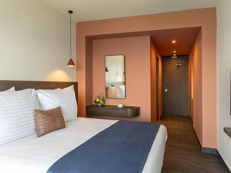 Modernes Hotelzimmer mit großem Bett, weißen und blauen Decken, warmen Wandfarben und minimalistischer Dekoration.