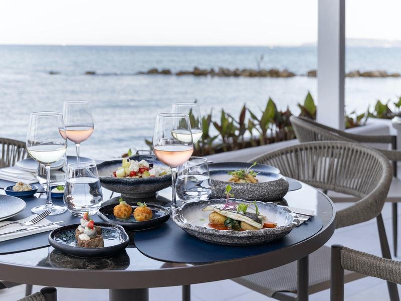 Eleganter gedeckter Tisch mit feinem Essen und Meerblick im Hintergrund, helle Stühle um den runden Tisch.