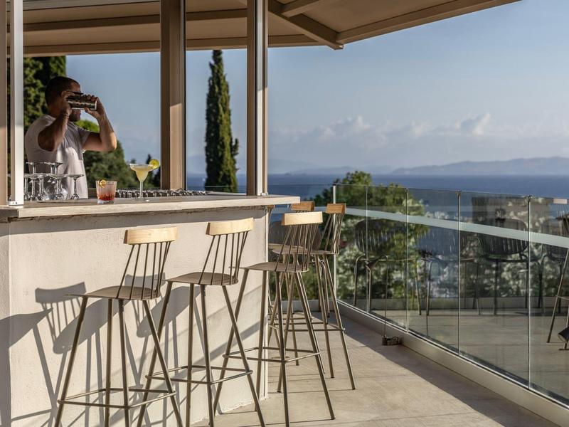 Offene Terrasse mit Bar, vier hohen Metallstühlen und Meerblick im Hintergrund bei klarem Himmel.