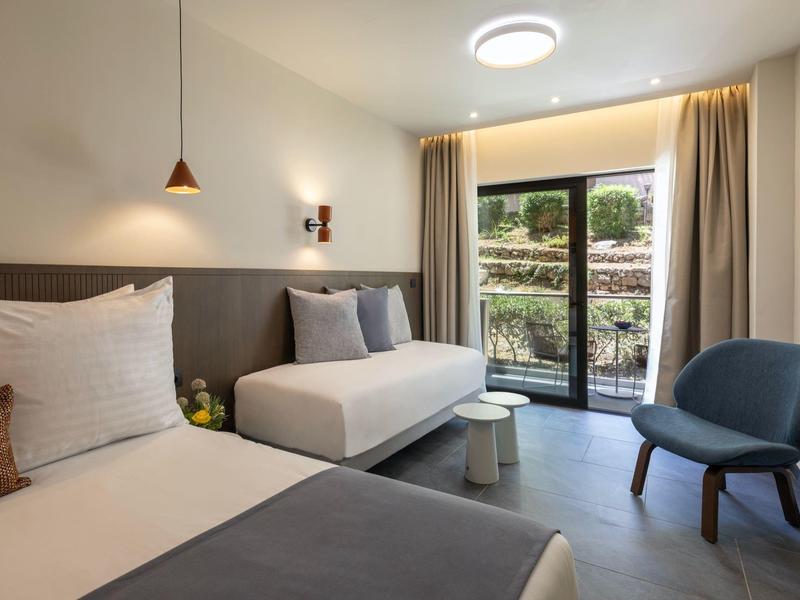 Hell eingerichtetes Zimmer mit zwei Betten, Stuhl, Nachttisch, Lampe und großem Fenster mit Vorhängen.