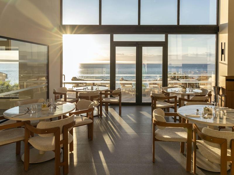 Helles Café mit Holzstühlen und Tischen, großen Fenstern und Meerblick bei Sonnenuntergang.