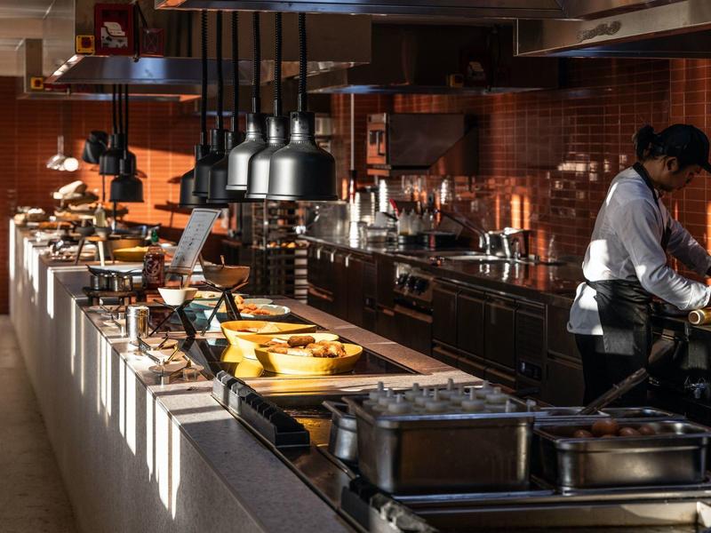 Moderne Profi-Küche mit Edelstahlgeräten, Holzregalen, warmem Licht und Koch in Arbeitskleidung.