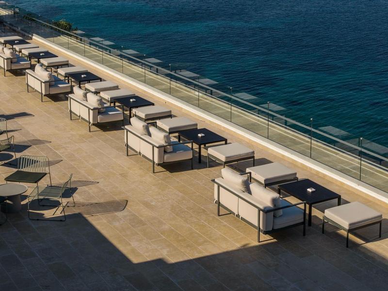 Reihe moderner Sitzgruppen mit Tischen an Terrasse neben blauem Meer unter sonnigem Himmel.