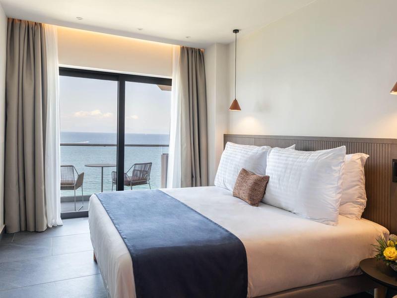 Modernes Schlafzimmer mit Doppelbett, großen Fenstern und Blick auf das Meer bei Sonnenuntergang.