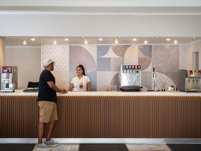 Recepción de hotel con un huésped y personal bajo luz cálida y patrón geométrico en la pared