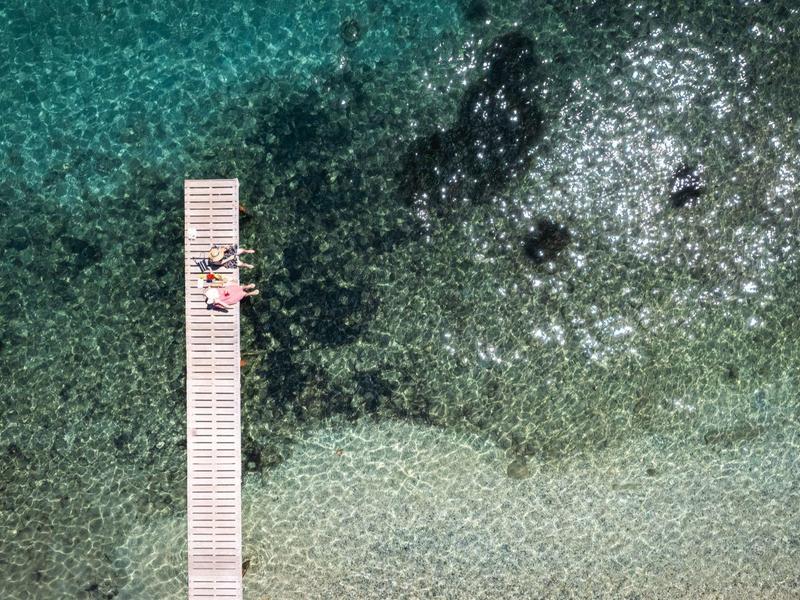 Ein Steg ragt ins klare, türkisblaue Wasser, zwei Personen liegen darauf und genießen die Sonne.