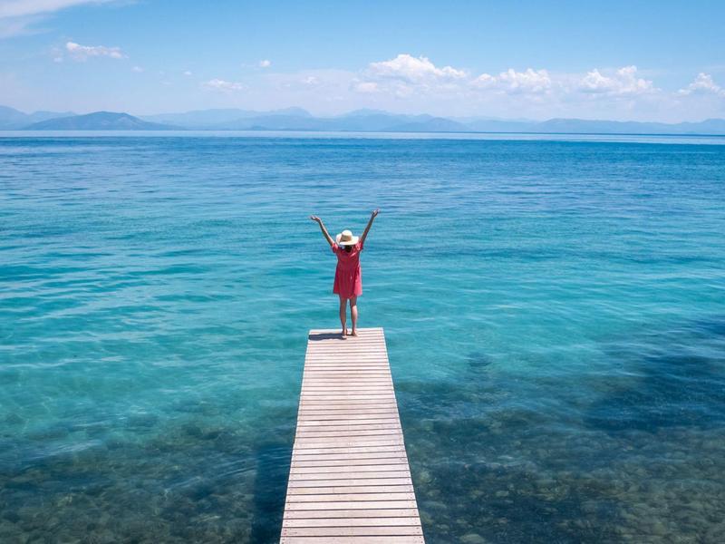 Ein Steg ragt ins klare, blaue Meer, darauf steht eine Person mit erhobenen Armen bei sonnigem Himmel.