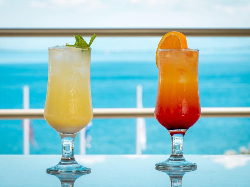Zwei bunte Cocktails mit Früchten stehen auf glattem Tisch vor blauem Meer und Himmel.