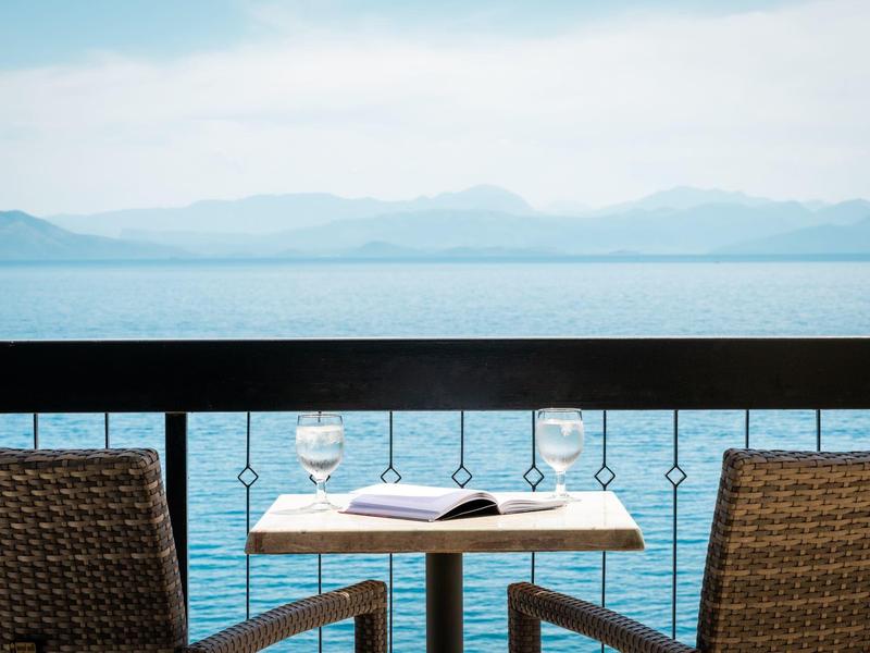 Zwei geflochtene Stühle am kleinen Tisch mit Buch und Glas, Blick auf ruhiges Meer und Berge.