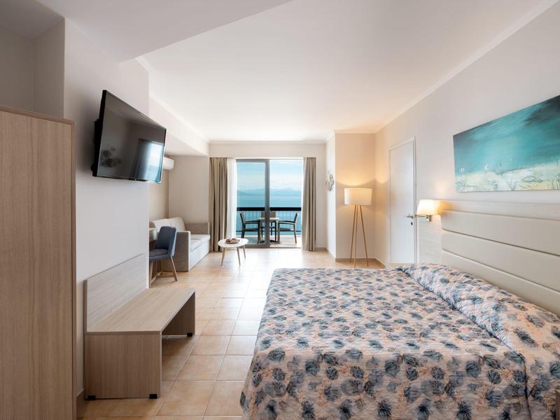 Helles Hotelzimmer mit Doppelbett, Fernseher, Schreibtisch, Balkon und Meerblick bei Tageslicht.