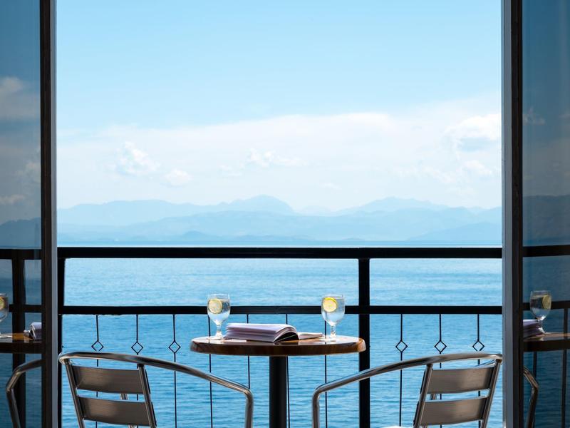 Balkontisch mit zwei Stühlen, Kerzen und Buch, Blick auf ruhiges Meer und Berge im Hintergrund.