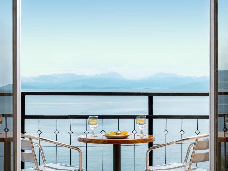 Balkontisch mit Getränken, Blick auf Meer und Berge bei klarem Himmel.