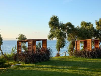 Zwei offene Holzpavillons in einem grünen Garten mit Blick aufs Meer bei klarem Himmel.