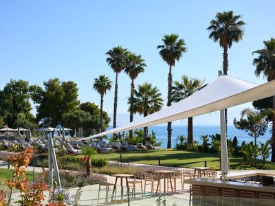 Moderner Außenbereich eines Hotels mit Palmen, Sitzgelegenheiten und Meerblick bei Sonnenschein.