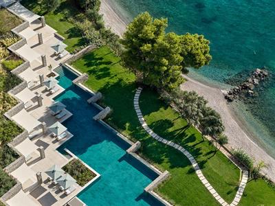 Vogelperspektive eines Hotels mit Pools, grünem Garten und angrenzendem Kiesstrand am Meer.