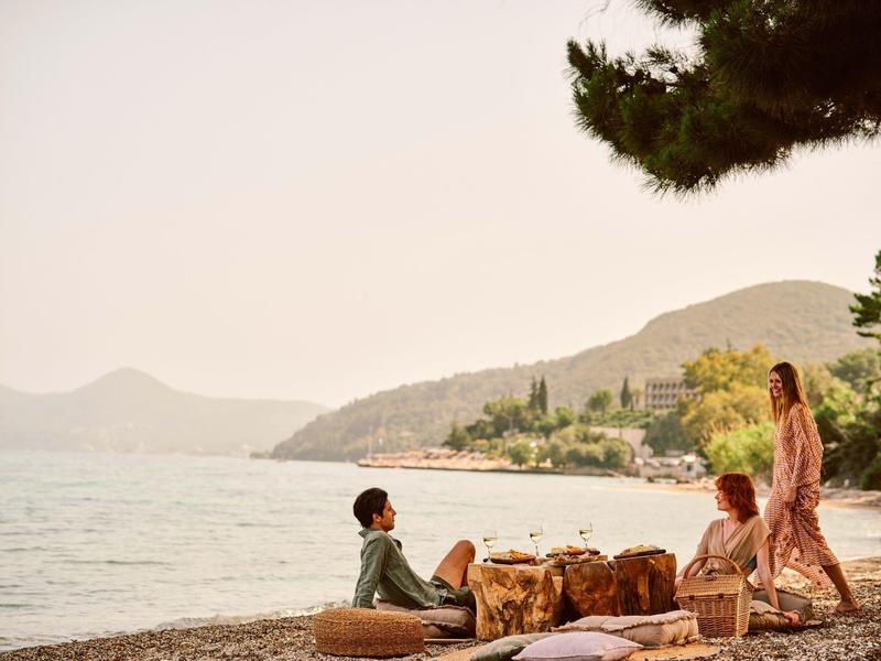 Drei Personen am Ufer, sitzend und stehend, mit Bergen, Wasser und Bäumen im Hintergrund.