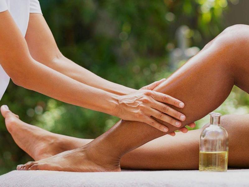Person massiert Bein einer anderen Person auf einer Liege, daneben Ölflasche, grüne Umgebung im Hintergrund.