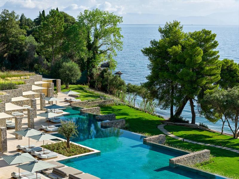 Moderner Poolbereich mit Sonnenschirmen, Liegestühlen und Blick auf das Meer in einer grünen Landschaft.