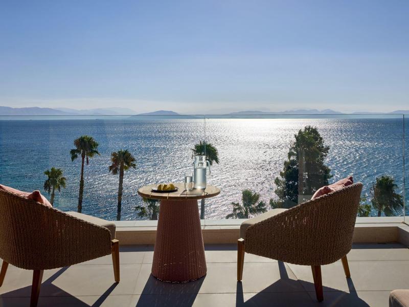 Zwei Stühle und ein Tisch auf einer Terrasse mit Meerblick und Palmen im Sonnenlicht.