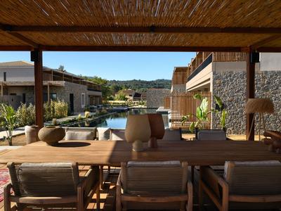 Überdachte Terrasse mit Holztisch und Stühlen, Blick auf einen Pool und Gebäude in sonniger Umgebung.