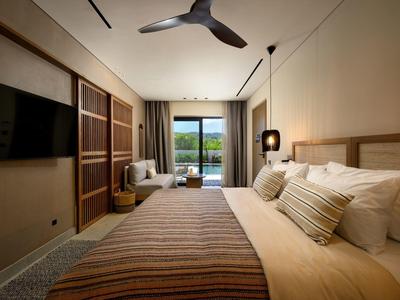 Schlafzimmer mit großem Bett, Fernseher, Deckenventilator und Blick auf Balkon mit grüner Landschaft.