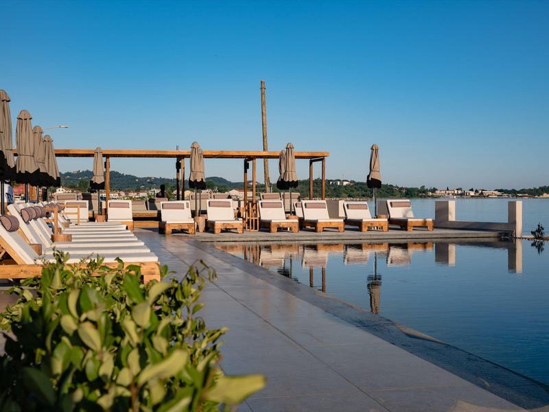 Poolbereich mit Liegen, Sonnenschirmen und klar blauem Himmel, umgeben von Wasser und Pflanzen.