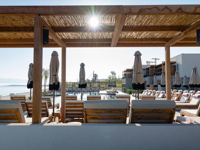 Strandbar mit Holzdächern, Sonnenschirmen, Liegestühlen und Blick auf das Meer im Hintergrund.