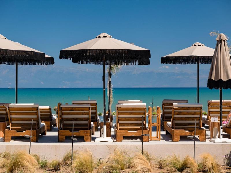 Strand mit mehreren Holzliegen und großen Sonnenschirmen vor blauem Meer und klarem Himmel.