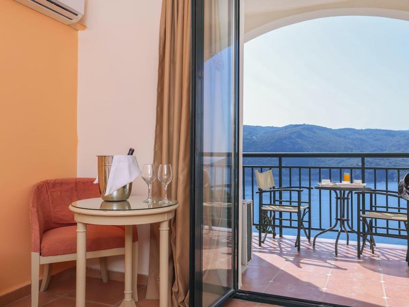 Balkon met tafel en twee stoelen met uitzicht op bergen, aangrenzend aan lichte kamer met tafel en fauteuil.