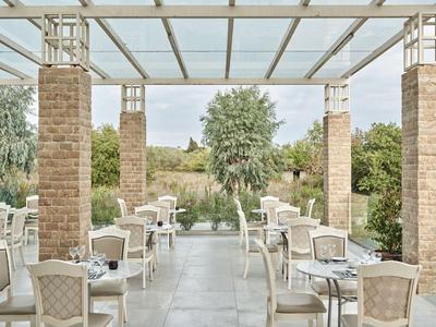 Terraza de restaurante con muebles blancos, pilares de ladrillo y vista a un jardín con árboles.